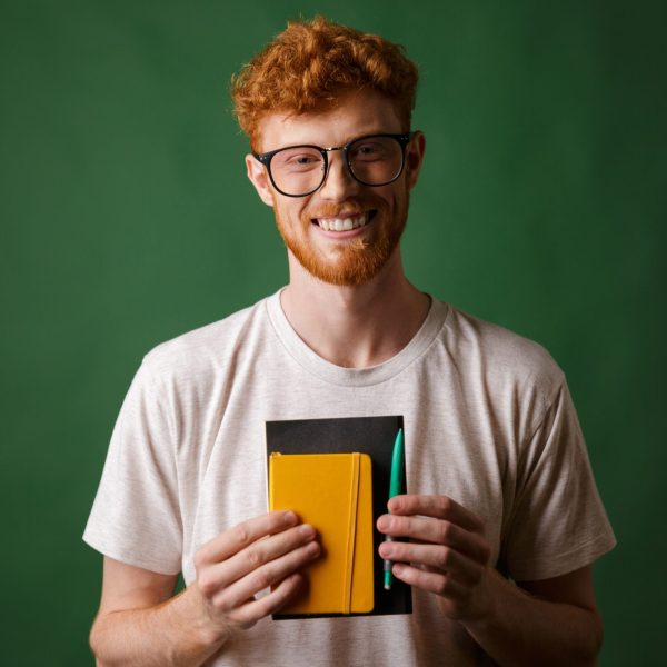 Portrait of smiling readhead bearded nerd, holding notebook and pen, looking at camera, over green background